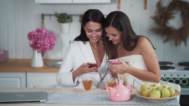 Dos mujeres desayunan, beben té, miran el teléfono y sonríen, compran en línea — Vídeo de stock