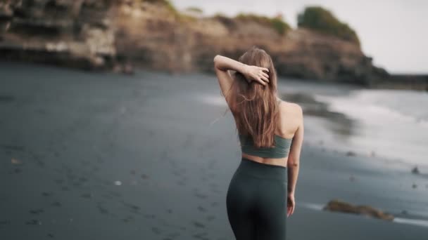 Backside view woman in sportswear walks along black sand beach and turns around — 비디오