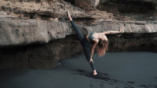 Young woman doing stretching on black sand beach near cliff, slow motion — Stock Video