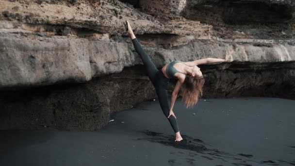 Jeune femme faisant des étirements sur la plage de sable noir près de la falaise, au ralenti — Video