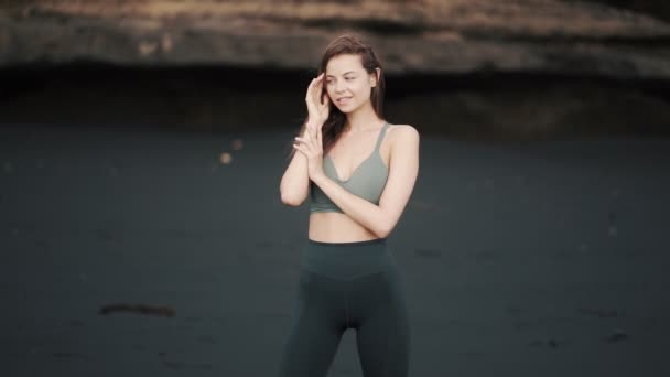 Mladá sexy žena ve sportovním oblečení pózuje na černé písečné pláži, dívá se na kameru, úsměvy