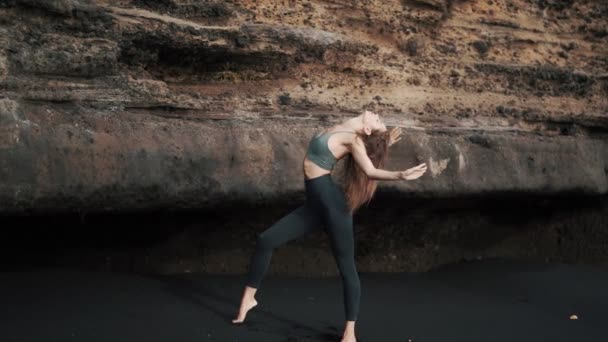 Женщина делает упражнения на растяжку на черном песке у скалы, замедленная съемка — стоковое видео
