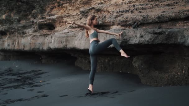 Kvinna gör stretching övningar på svart sandstrand nära klippan, slow motion — Stockvideo