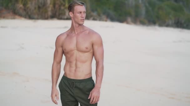 Hezký muž bez trička se svalnatým tělem stojí na pláži a dívá se na oceán — Stock video