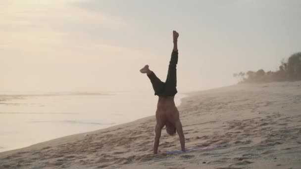 Man gör headstand under träning på stranden, står på armarna, sedan sitter på tvinnat garn — Stockvideo