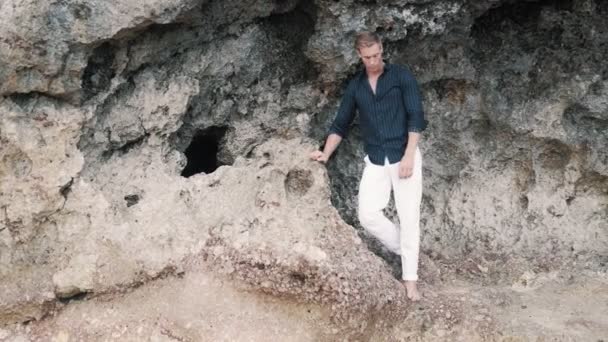 Homem brutal em calças brancas e camisa preta fica na praia perto da rocha, olha para longe — Vídeo de Stock