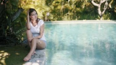 Kadın havuzun kenarında oturuyor, suya eliyle dokunuyor ve su damlatıyor.