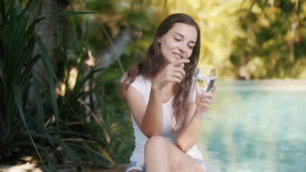 Porträtt av kvinna sitter på kanten av poolen, håller glas vatten i handen, ler — Stockvideo