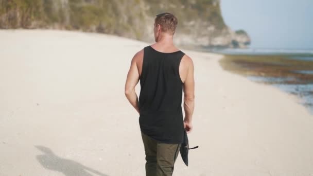 Вид ззаду, самотній чоловік з йоги килимок в руках ходить піщаним пляжем — стокове відео