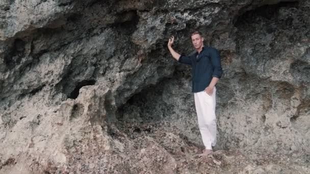 Brutaler Mann in weißer Hose und schwarzem Hemd steht am Strand in der Nähe von Felsen, schaut weg — Stockvideo