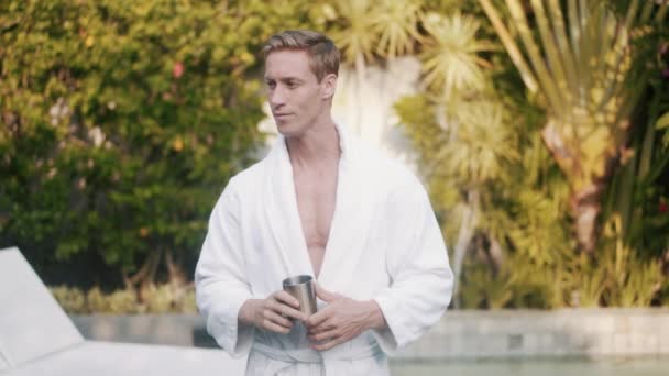 Man i badrock dricker kaffe tidigt på morgonen utomhus, grönska på bakgrunden — Stockvideo