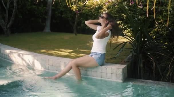 Mujer en gafas de sol y ropa casual se sienta en el borde de la piscina, salpica el agua con los pies — Vídeos de Stock
