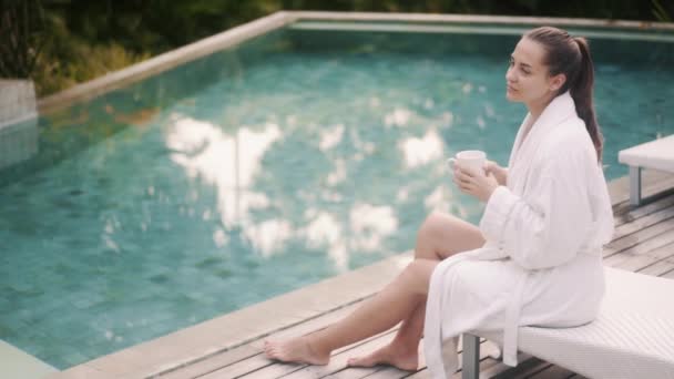 Mujer en albornoz blanco se sienta en la cama de sol cerca de la piscina, bebe café por la mañana temprano — Vídeos de Stock