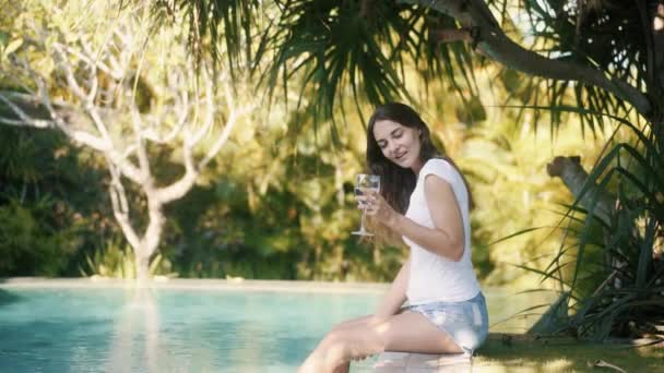 Vrouw zit aan de rand van het zwembad, spat water met voeten en drinkt water uit glas — Stockvideo