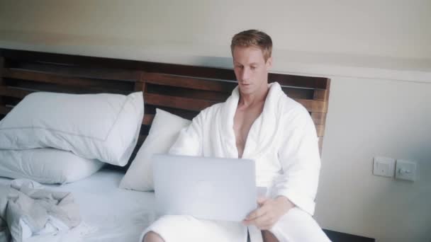Bonito homem de roupão branco fica na cama em seu apartamento e usa laptop para o trabalho — Vídeo de Stock