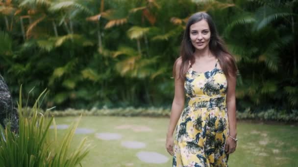 Žena kráčí tropickou zahradou ke kameře, dotýká se rukou vlasů, usmívá se — Stock video
