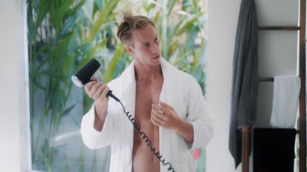 Beyaz bornozlu, sportif vücutlu bir adam saç kurutma makinesiyle saçlarını kurutuyor. — Stok video