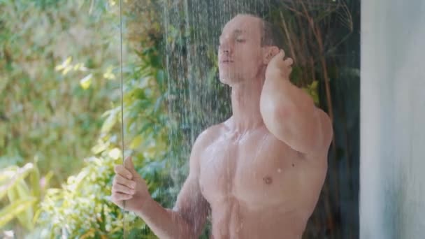 Sexy man with muscular body takes shower, tropical greenery on background — Stock Video