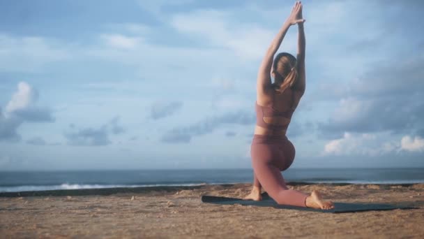 Vrouw doet yoga oefeningen en strekt zich 's morgens op mat op het strand, slow motion — Stockvideo