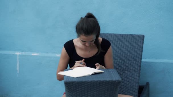 Giovane donna si siede sul lettino, prende appunti nel diario, utilizza cellulare, sfondo blu — Video Stock