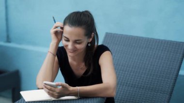 Young woman sits on sunbed, makes notes in diary, uses mobile, blue background