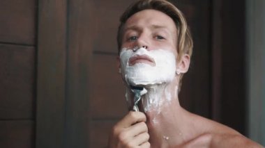 Portrait of man looking at camera and shaving his beard with manual razor
