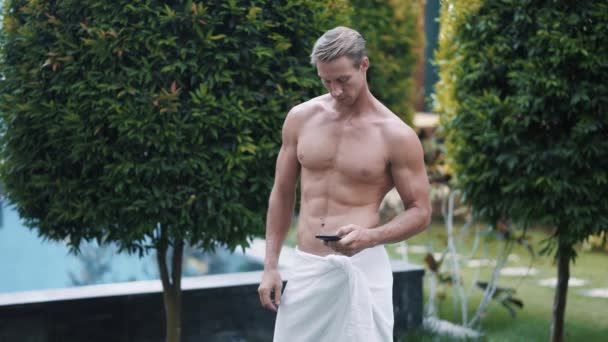 Portrait of sexy muscular shirtless man uses mobile, greenery on background — Stock video