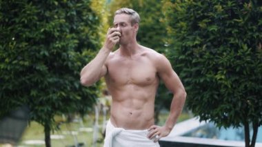 Portrait of sexy muscular shirtless man drinks coffee, greenery on background