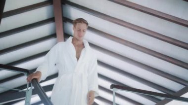 handsome man in white bathrobe goes down stairs in house
