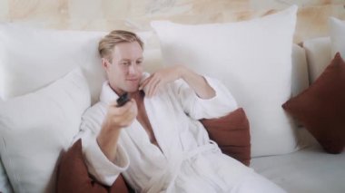 blond guy in bathrobe switches on TV on comfortable bed
