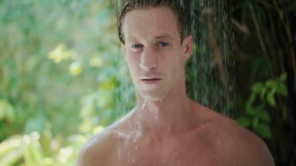 Young man takes refreshing shower near green tropical plants — Stockvideo