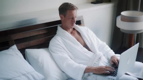 Приваблива людина в халаті працює на ноутбуці в зручному ліжку — стокове відео