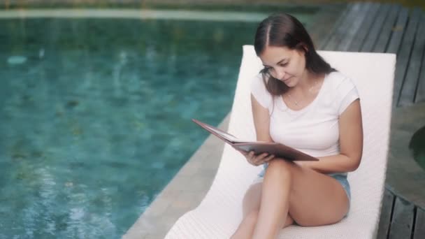 Beautiful girl chooses dish in menu near swimming pool — 图库视频影像