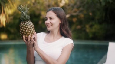 cheerful woman holds ripe pineapple against swimming pool