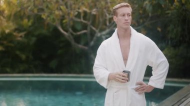 young man in bathrobe drinks coffee near swimming pool