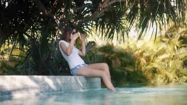 Girl in t-shirt and denim shorts swings feet in pool water — Wideo stockowe