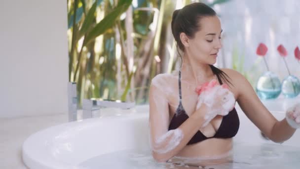 Дівчина в бікіні миє тіло, приймаючи ванну кімнату з рослинами — стокове відео