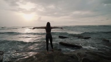 girl stands on stone and enjoys sun setting down above ocean