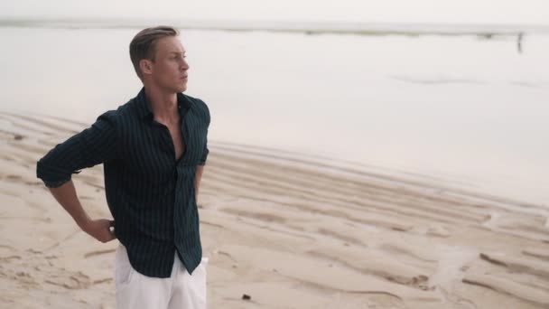 Blue eyes blond guy poses on empty beach near volcanic rocks — стоковое видео