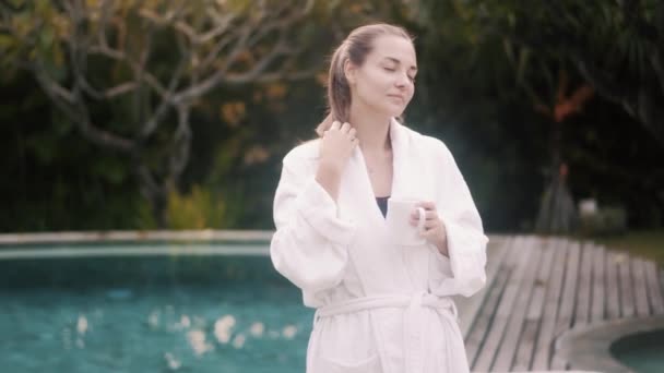 Beautiful girl in bathrobe drinks coffee near swimming pool — Αρχείο Βίντεο