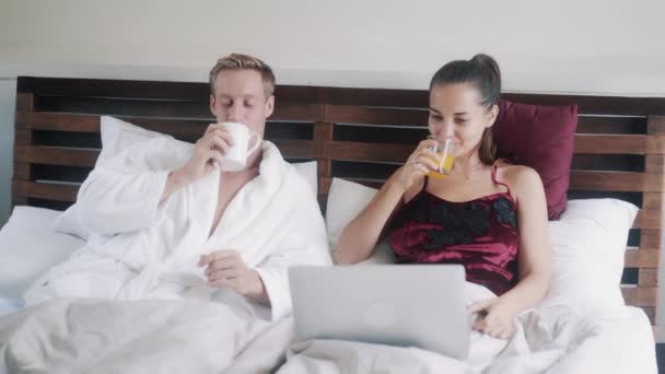 Beautiful couple with laptop drinks juice resting in bed — Stock video
