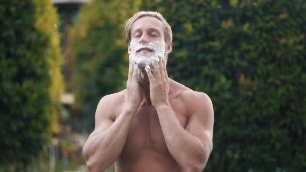Счастливый человек применяет пену для бритья на лице в зеленом саду — стоковое видео