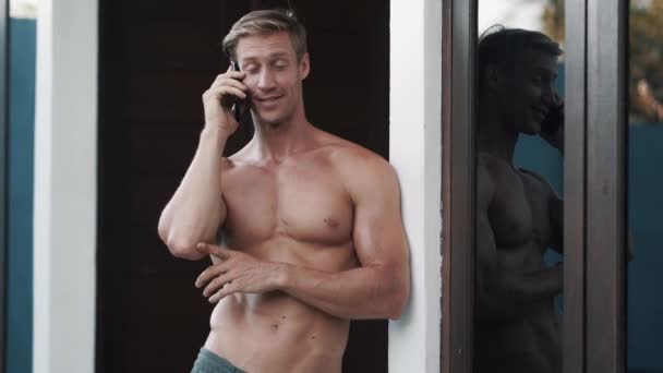 Молодой человек разговаривает по смартфону через стеклянную дверь дома — стоковое видео