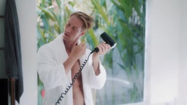 young man dries blond hair near large mirror in bathroom
