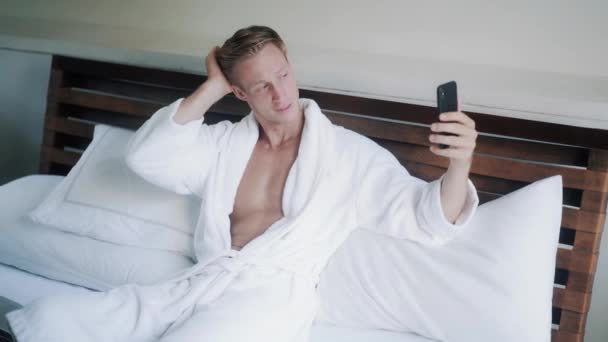 Guapo rubio chico utiliza videochat en teléfono móvil en la cama — Vídeo de stock