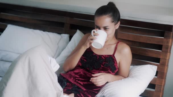 Doamnă în pijama bea cafea întinsă pe pat mare dimineața — Videoclip de stoc