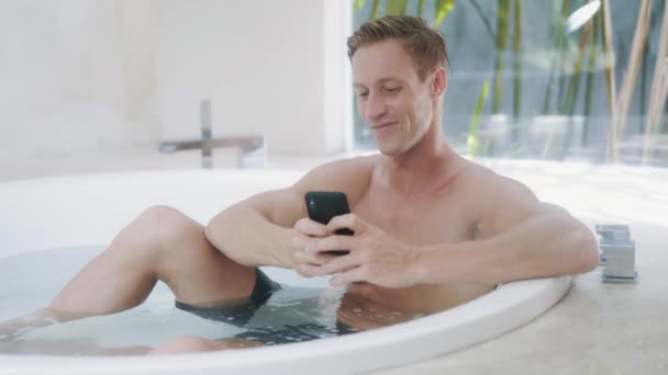 Young man reads messages on smartphone in modern bathtub — ストック動画