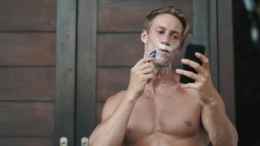blond man shaves face with razor looking at phone near door