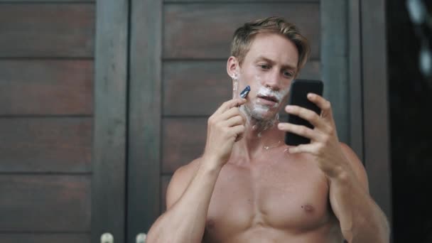 Blond man shaves face with razor looking at phone near door — Stockvideo