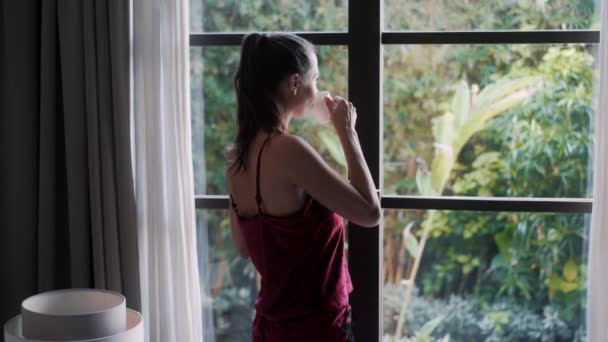 Vista lateral da mulher de pijama bebendo café de manhã e olhando pela janela — Vídeo de Stock